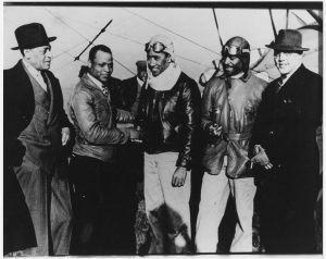 Dale L. White (center) and Chauncey E. Spencer (second from right) are greeted by National Airmen's Association President Cornelius R. Coffey (second from left) at Harlem Airport, Chicago. Chicago businessman James Hill (far left) and author and sociologist Horace C. Cayton (far right) also appear.