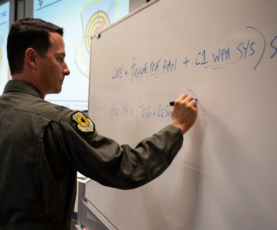 Col. Kevin Jamieson, 57th Wing vice commander, writes on a white board at Nellis Air Force Base, Nev., July 2021. Jamieson provided graduates from class 21-B of the U.S. Air Force Advanced Maintenance and Munitions Operations School with equations on how to successfully acquire mission capabilities and effective operations. (Air Force photograph by Senior Airman Bryan Guthrie)