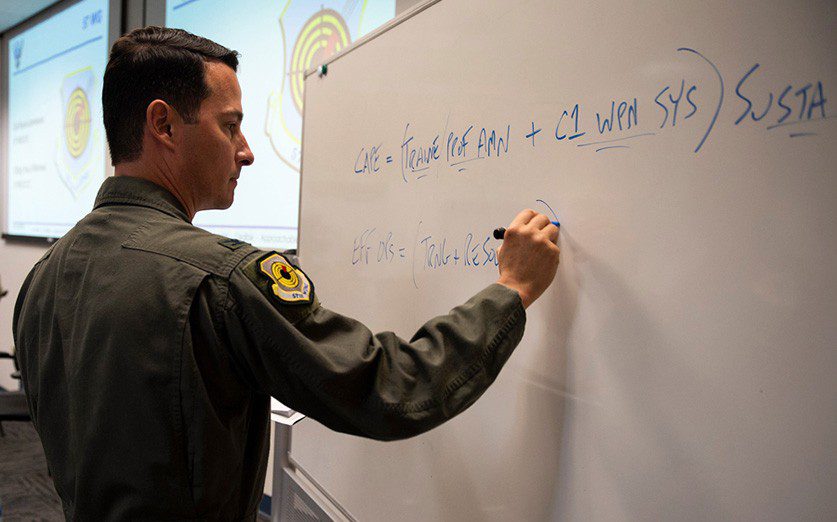 Col. Kevin Jamieson, 57th Wing vice commander, writes on a white board at Nellis Air Force Base, Nev., July 2021. Jamieson provided graduates from class 21-B of the U.S. Air Force Advanced Maintenance and Munitions Operations School with equations on how to successfully acquire mission capabilities and effective operations. (Air Force photograph by Senior Airman Bryan Guthrie)