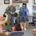Photograph by Linda KC Reynolds Stephanie Cregier, director of the Junior Test Pilot School Summer Museum Program, dons flight gear with a little help from B-2 Test Pilot Don Weiss. Weiss also showed students what pilots must wear in order to be safe at high altitudes and how to survive when ejecting from an aircraft and landing in water.