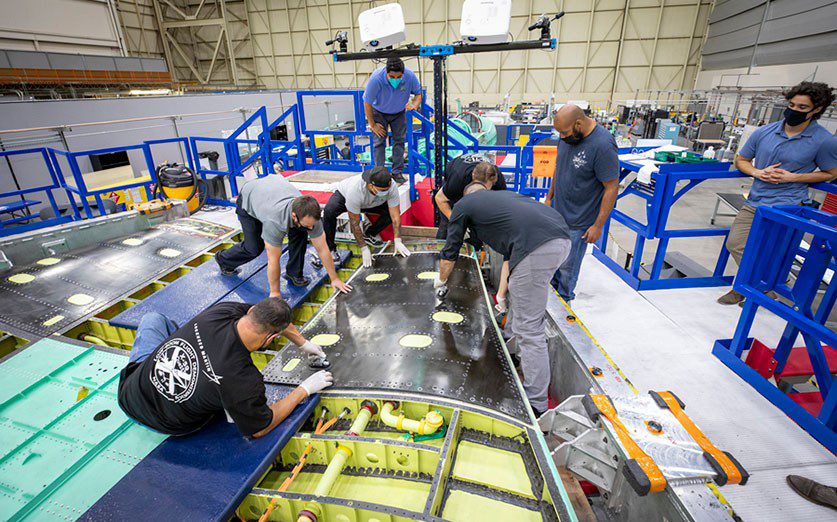 A panel of the dark-colored skin of the wing of NASA’s X-59 Quiet SuperSonic Technology airplane is laid in place over the wing’s interior framework for permanent attachment. The volume underneath will serve as a fuel tank. The X-59 is under construction by Lockheed Martin at the company’s Skunk Works factory in California. (Lockheed Martin photograph)